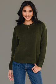 A woman with long dark hair smiles while wearing the Panache Apparel Collection Dark Green Soft Sweater, paired with blue jeans and gold jewelry, as she stands against a plain gray background.