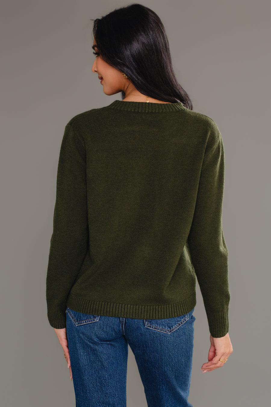 A person with long dark hair models the Panache Apparel Collection's Dark Green Soft Sweater, paired with blue jeans, standing facing away from the camera against a plain gray background.