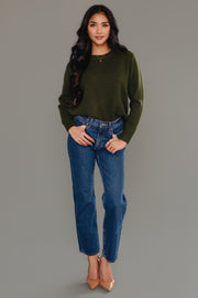 A woman with long dark hair wears the Panache Apparel Collection Dark Green Soft Sweater, styled with blue jeans and tan high heels, as she stands against a plain gray background, smiling with one hand in her pocket.