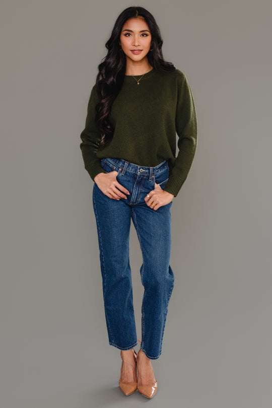 A woman with long dark hair wears the Panache Apparel Collection Dark Green Soft Sweater, styled with blue jeans and tan high heels, as she stands against a plain gray background, smiling with one hand in her pocket.