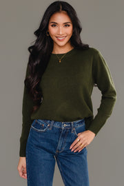 A woman with long black hair is smiling with one hand in her pocket, wearing the Panache Apparel Collection Dark Green Soft Sweater and blue jeans, standing against a plain gray background.