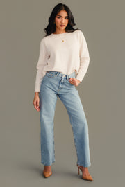 A woman poses against a plain taupe background wearing the Panache Apparel Collection Cream Soft Sweater, paired with light blue high-waisted jeans and brown high-heeled shoes. She stands with one hand on her hip while the other rests relaxed at her side.