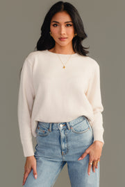 A woman with long dark hair models the Panache Apparel Collection Cream Soft Sweater, paired with light blue jeans, standing with one hand in her pocket against a plain gray background.