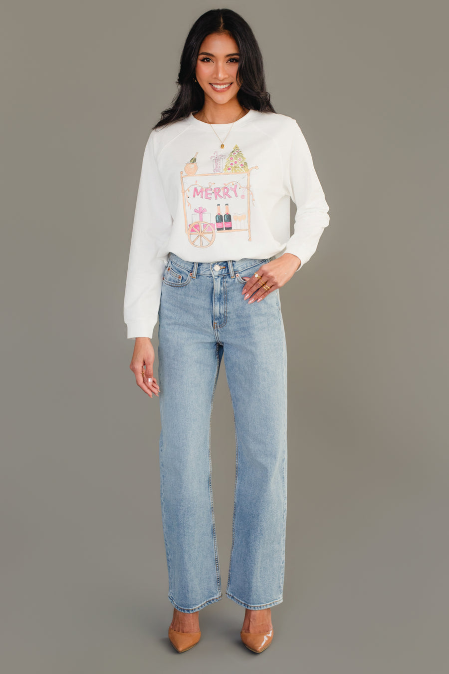 A woman wearing a white long-sleeve Merry Bar Cart Sweatshirt by Panache Apparel Collection, featuring a festive bar cart graphic, paired with light-wash straight-leg jeans.