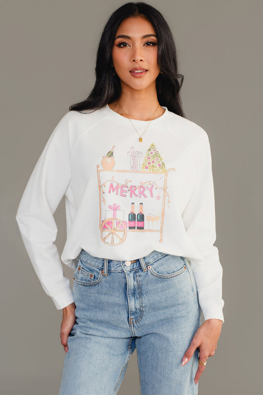A white Merry Bar Cart Sweatshirt featuring a graphic of a holiday-decorated bar cart with the word MERRY hanging from it, worn with light-wash jeans.