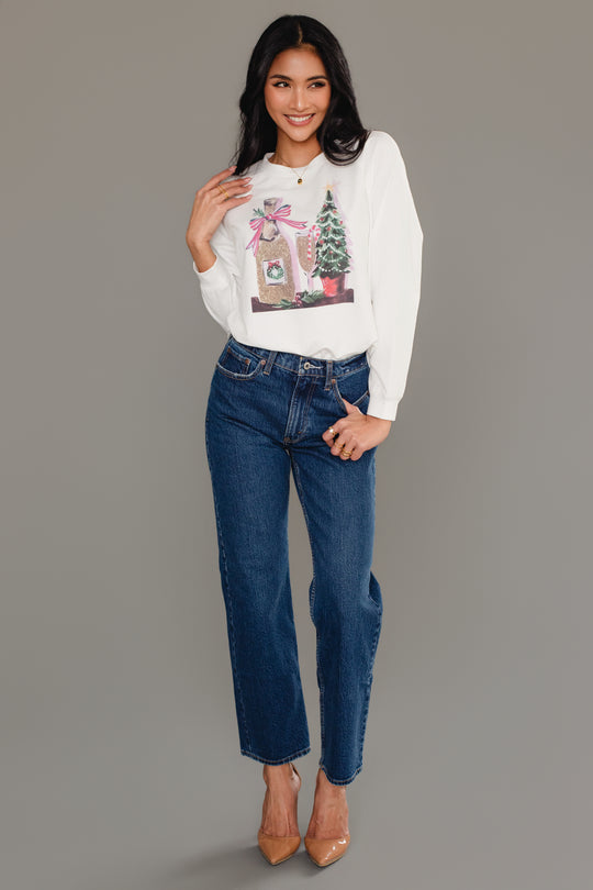 A woman wearing a white Christmas Champagne Sweatshirt by Panache Apparel Collection with a champagne bottle and Christmas tree graphic, paired with blue jeans and tan heels.