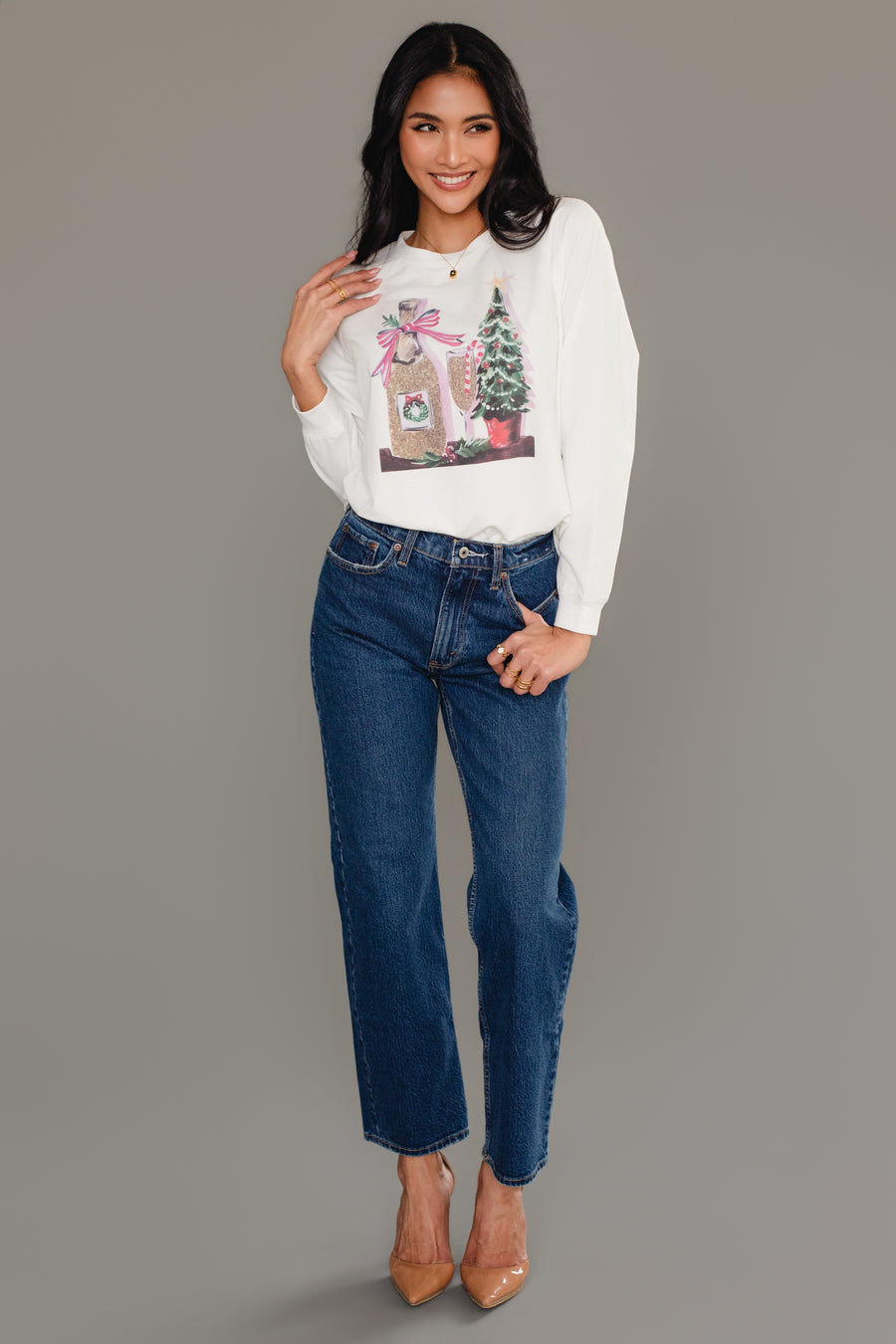 A woman wearing a white Christmas Champagne Sweatshirt by Panache Apparel Collection with a champagne bottle and Christmas tree graphic, paired with blue jeans and tan heels.