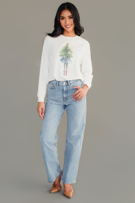 A woman wearing a Girl With Christmas Tree Sweatshirt in white, paired with light-wash jeans and tan heels.