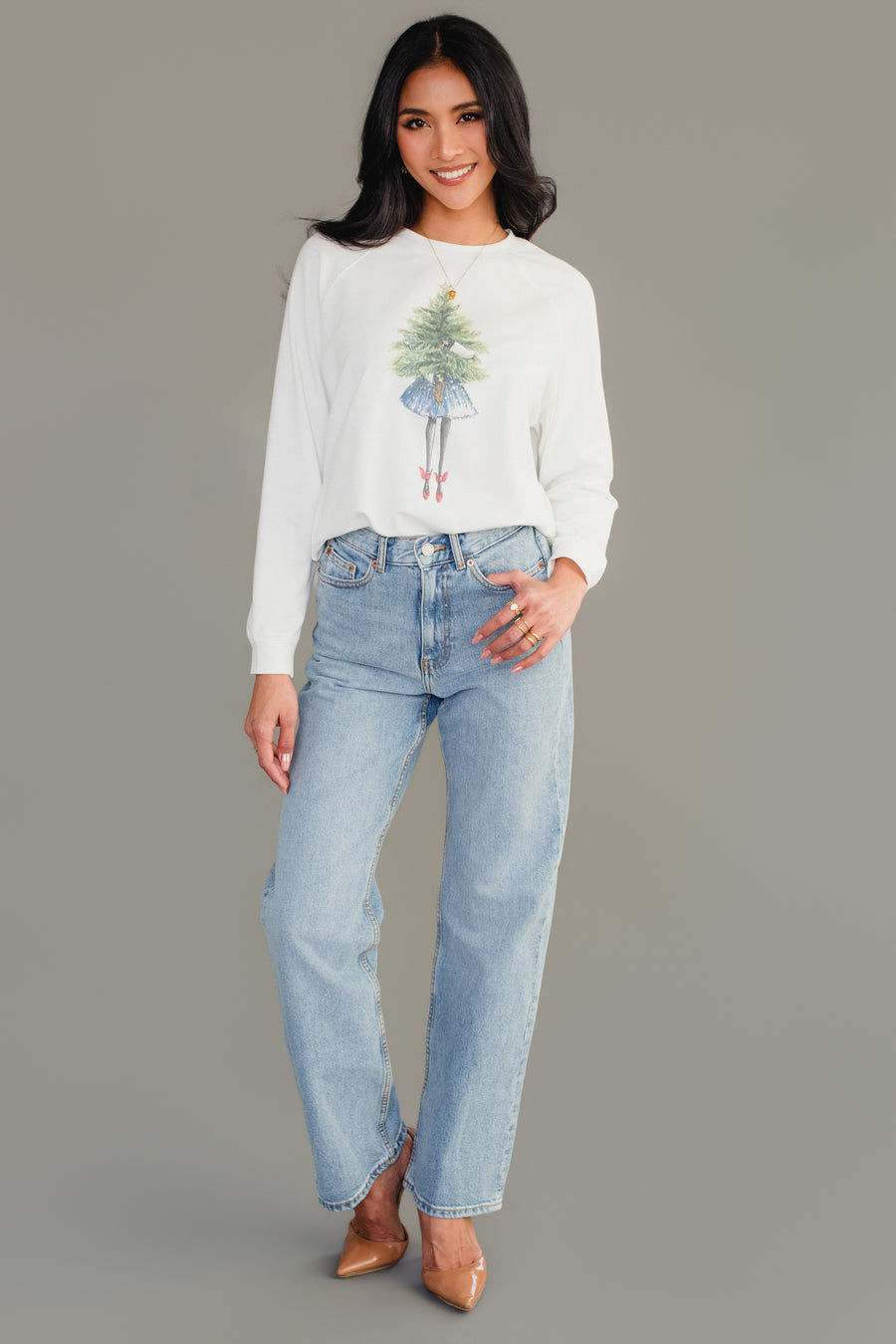 A woman wearing a Girl With Christmas Tree Sweatshirt in white, paired with light-wash jeans and tan heels.