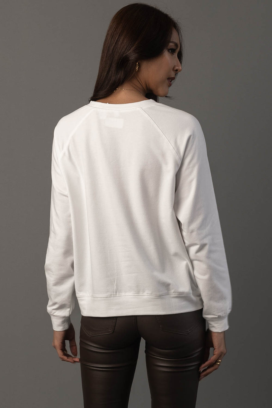 A woman with long brown hair, wearing the Panache Apparel Collection’s Girl At Pumpkin Patch Sweatshirt and dark pants, stands with her back to the camera against a plain gray background.