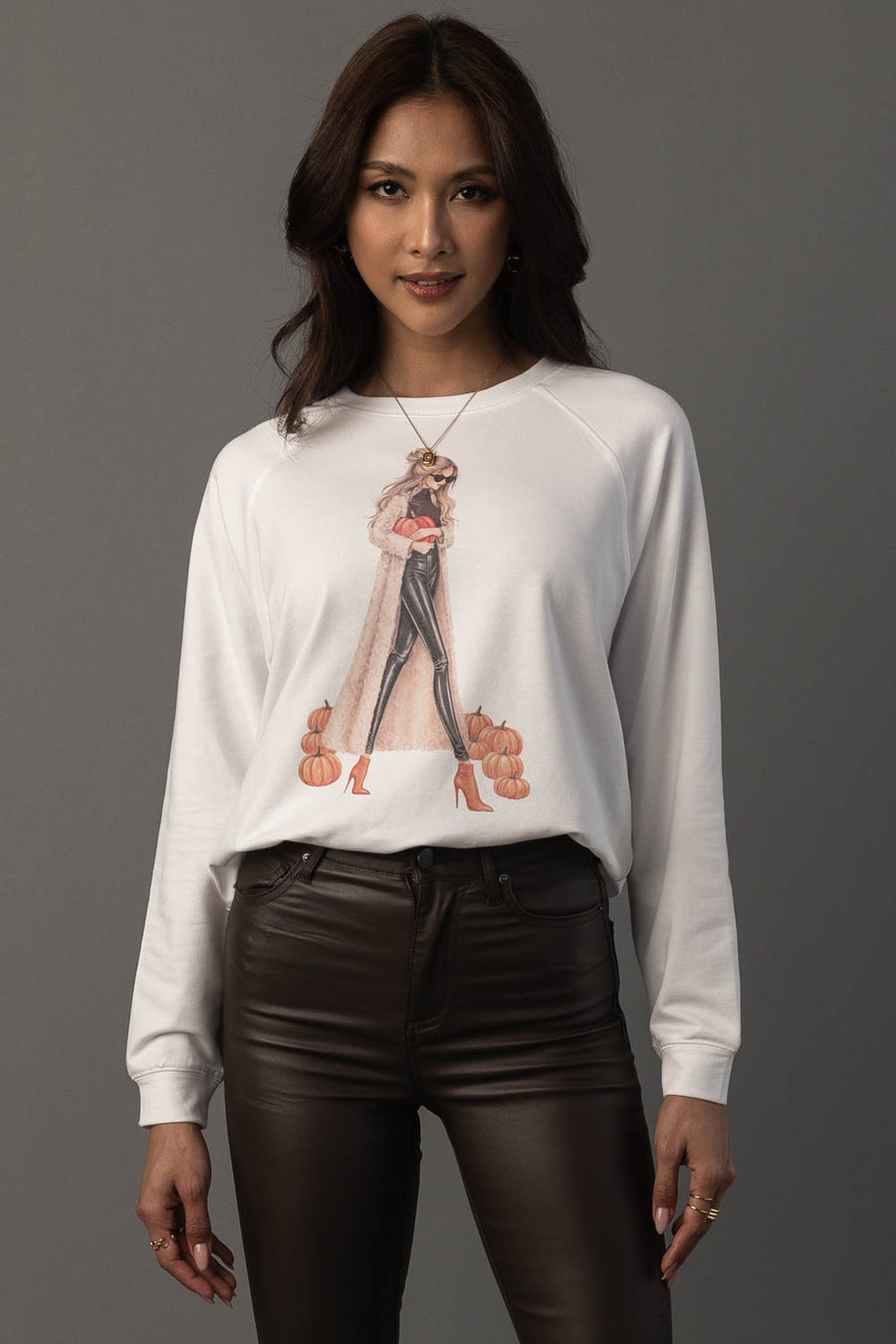 A woman stands against a gray background wearing the “Girl At Pumpkin Patch Sweatshirt” by Panache Apparel Collection, featuring a fashion illustration, paired with black leather pants. She has long dark hair and a neutral expression.