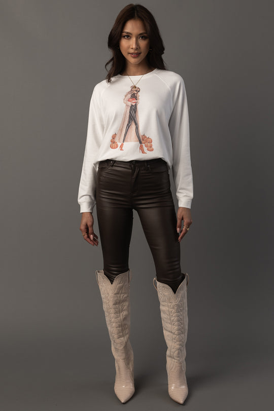 A woman stands against a plain background wearing the Panache Apparel Collection’s “Girl At Pumpkin Patch” sweatshirt, paired with black leather pants and light-colored knee-high cowboy boots.