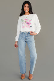 A woman wears a white Santa Bar Cart Sweatshirt featuring a graphic of Santa Claus at a bar cart, paired with light-wash jeans and tan heels.