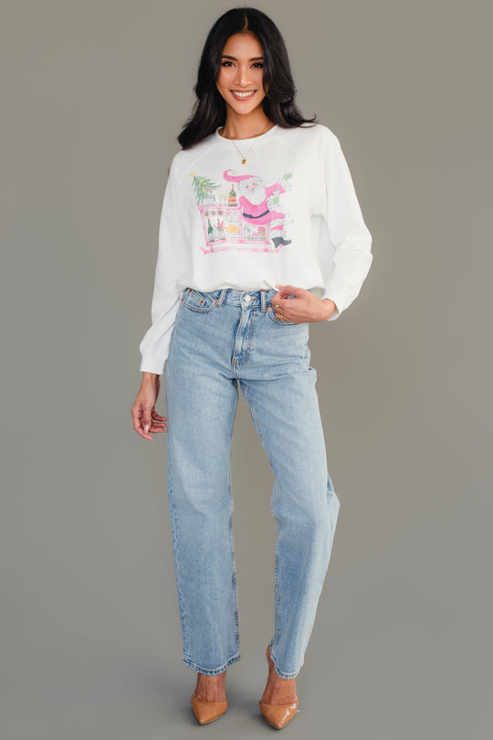 A woman wears a white Santa Bar Cart Sweatshirt featuring a graphic of Santa Claus at a bar cart, paired with light-wash jeans and tan heels.