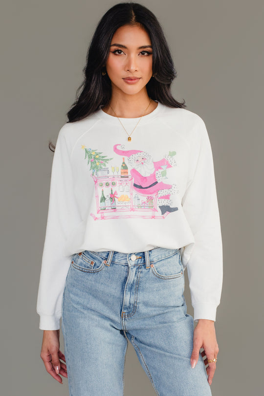 A person wears a white long-sleeve Santa Bar Cart Sweatshirt by Panache Apparel Collection featuring an illustration of Santa Claus standing at a bar cart with drinks and a small Christmas tree.