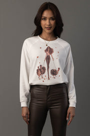 A woman with long brown hair wears the Panache Apparel Collection’s “Girl Walking Turkeys Sweatshirt,” which features an illustration of a woman walking two turkeys. She pairs it with dark brown pants and stands against a plain gray background.
