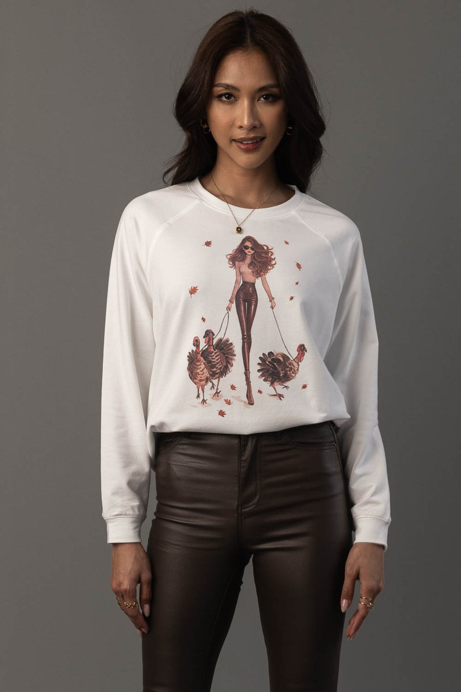 A woman with long brown hair wears the Panache Apparel Collection’s “Girl Walking Turkeys Sweatshirt,” which features an illustration of a woman walking two turkeys. She pairs it with dark brown pants and stands against a plain gray background.