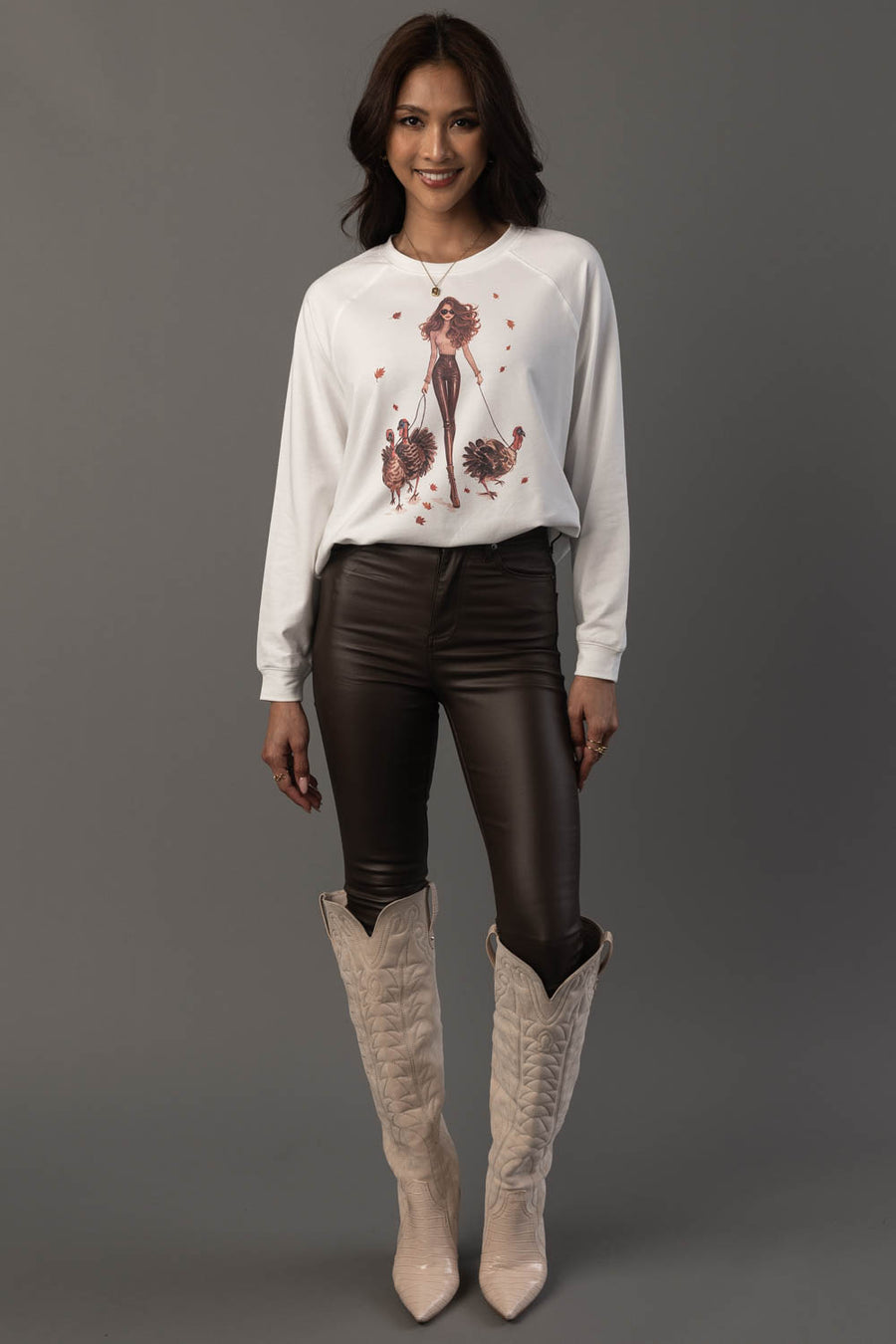 A woman with long dark hair stands and smiles against a plain gray background, wearing the Panache Apparel Collection’s Girl Walking Turkeys Sweatshirt in white, paired with dark leather pants and knee-high beige cowboy boots.