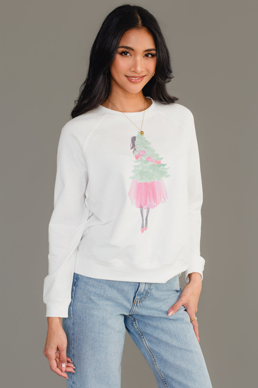 A person wearing a white long-sleeve Pink Girl Holding Tree Sweatshirt from the Panache Apparel Collection featuring a graphic of a girl in a pink skirt holding a Christmas tree.