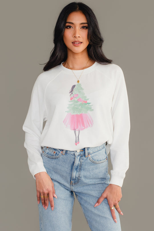 A person wears a white long-sleeve Pink Girl Holding Tree Sweatshirt featuring an illustration of a girl in a pink skirt hugging a Christmas tree.