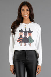 A woman smiles while wearing the Panache Apparel Collection’s Halloween Girls Sweatshirt, a white top featuring a graphic of three witches in hats and dresses, paired with black pants, standing against a plain gray background.