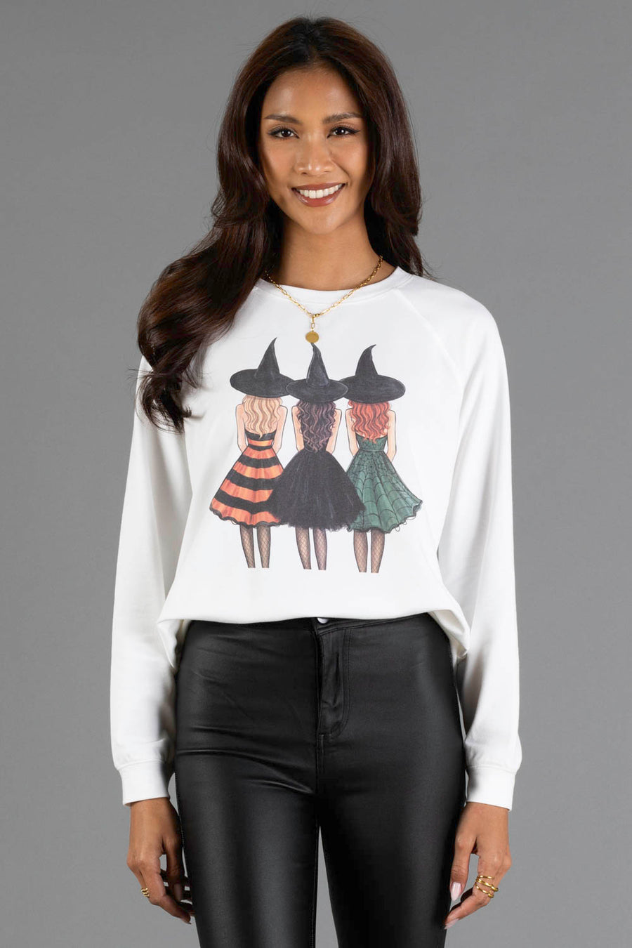 A woman smiles while wearing the Panache Apparel Collection’s Halloween Girls Sweatshirt, a white top featuring a graphic of three witches in hats and dresses, paired with black pants, standing against a plain gray background.