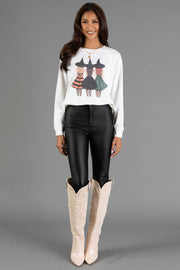 A woman stands in front of a gray background wearing the Halloween Girls Sweatshirt by Panache Apparel Collection, paired with black pants and tall cream-colored cowboy boots.