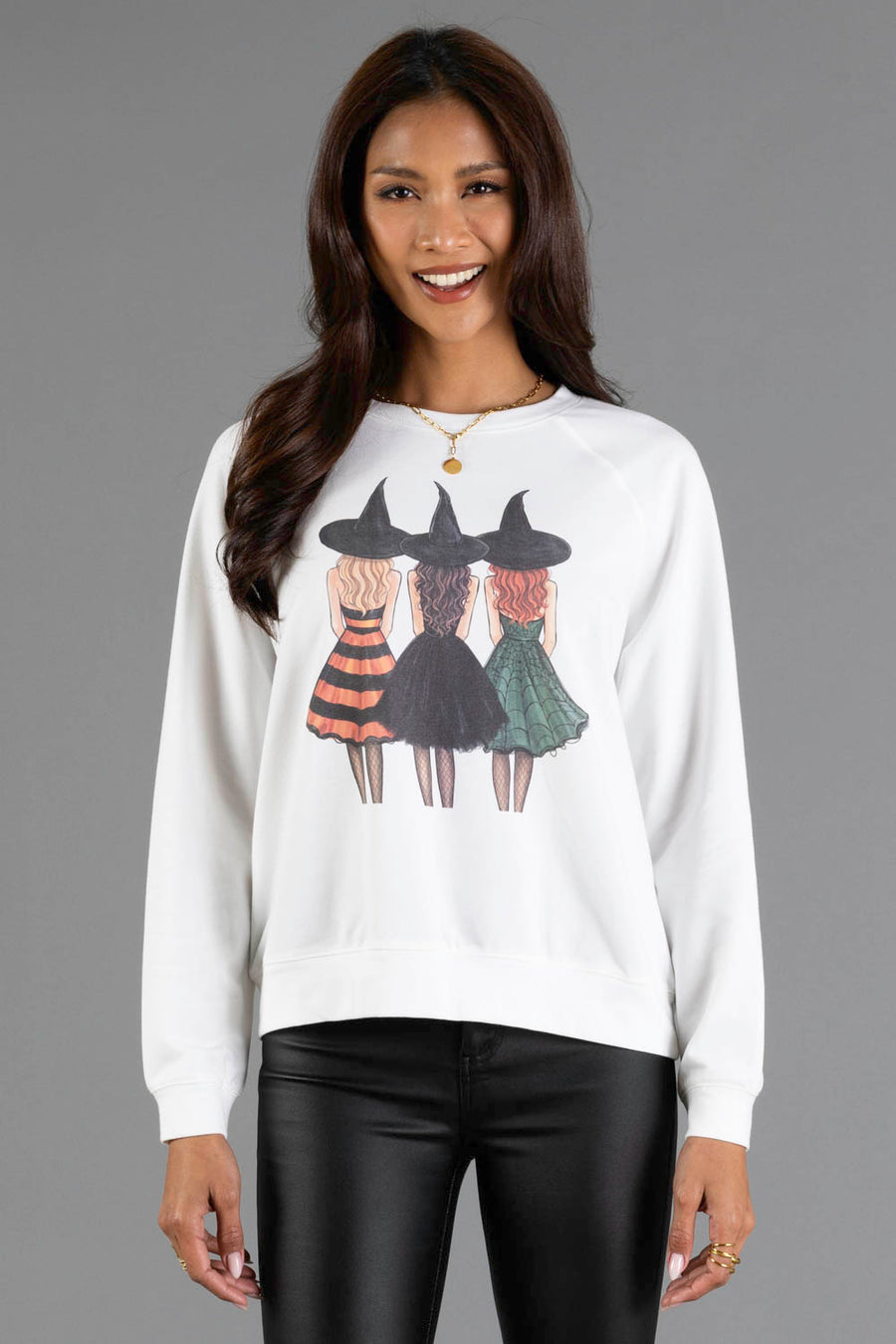 A woman with long dark hair and a smile wears the Panache Apparel Collection “Halloween Girls Sweatshirt,” featuring an illustration of three witches in colorful dresses and hats standing together. She pairs it with black pants.