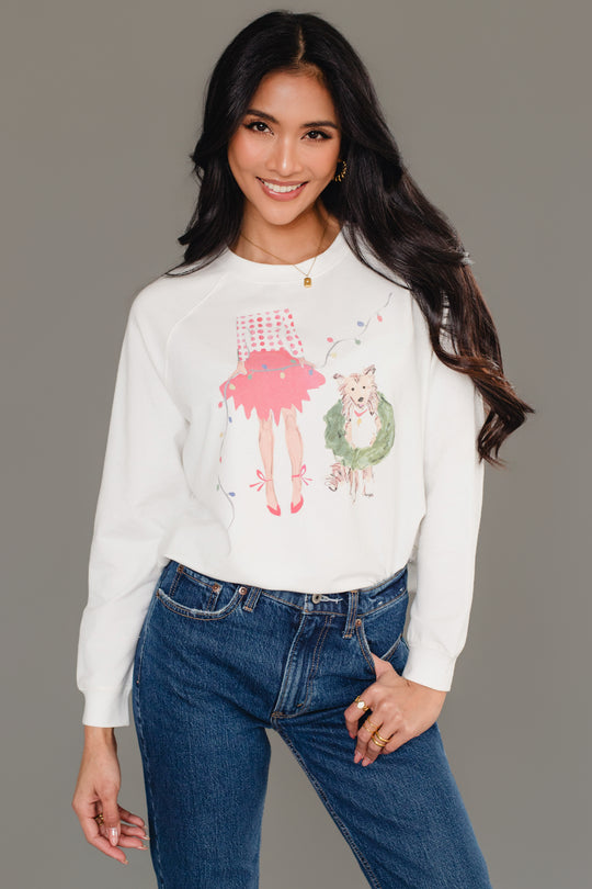A woman wears a white Christmas Girl & Dog Sweatshirt featuring a watercolor-style illustration of a girl in a red skirt and a dog wearing a wreath.