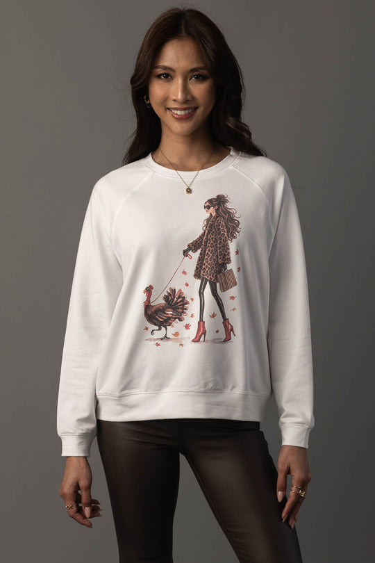 A woman models the Panache Apparel Collection’s “Girl Walking a Turkey Sweatshirt,” featuring a chic illustration of a stylish woman walking a turkey. She pairs it with dark pants and smiles against a plain gray background.