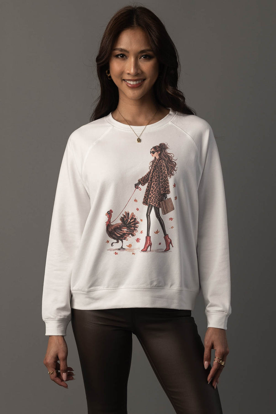 A woman models the Panache Apparel Collection’s “Girl Walking a Turkey Sweatshirt,” featuring a chic illustration of a stylish woman walking a turkey. She pairs it with dark pants and smiles against a plain gray background.