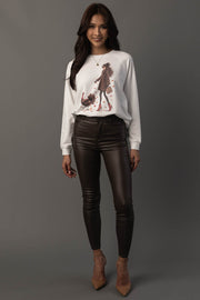 A woman, smiling at the camera, stands against a gray background wearing Panache Apparel Collection’s “Girl Walking a Turkey” Sweatshirt, paired with black leather pants and tan heels.