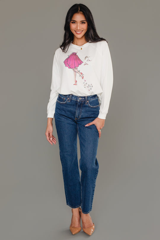 A woman wearing a white long-sleeve Fancy Christmas Fa La La Sweatshirt with a graphic of a pink skirt and the words fa la la, paired with dark blue jeans and tan heels.
