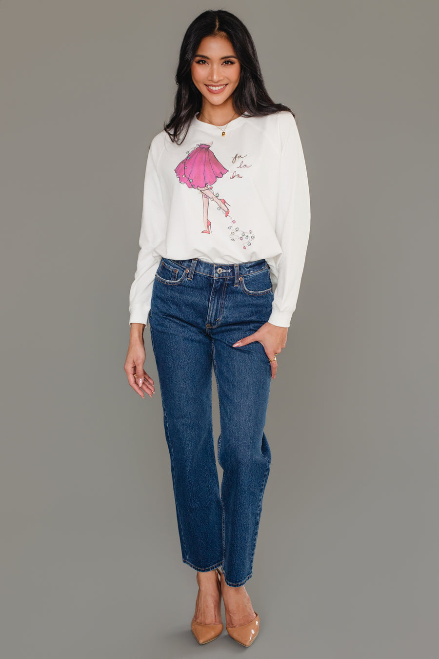 A woman wearing a white long-sleeve Fancy Christmas Fa La La Sweatshirt with a graphic of a pink skirt and the words fa la la, paired with dark blue jeans and tan heels.