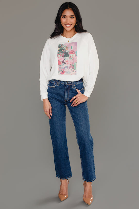 A smiling woman wears a white long-sleeve Christmas Cocktails Scene Sweatshirt featuring a graphic of cocktails and presents, paired with blue jeans and tan heels.