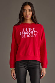 A person wears a red Tis The Season To Be Jolly Sweatshirt with the words printed in light pink.