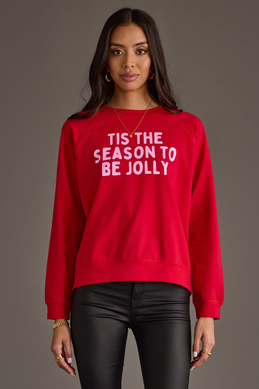 A person wears a red Tis The Season To Be Jolly Sweatshirt with the words printed in light pink.