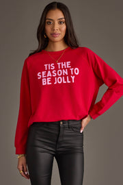 A person wears a red Tis The Season To Be Jolly Sweatshirt with pink lettering, paired with black pants.