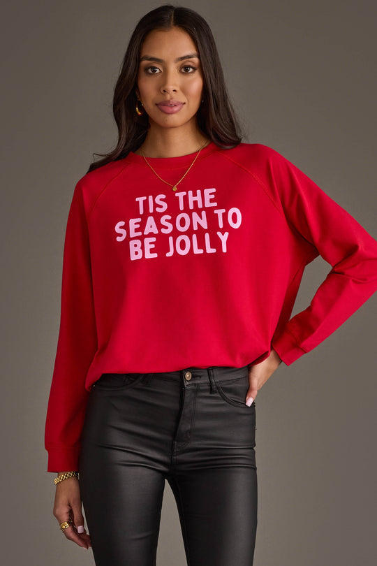 A person wears a red Tis The Season To Be Jolly Sweatshirt with pink lettering, paired with black pants.