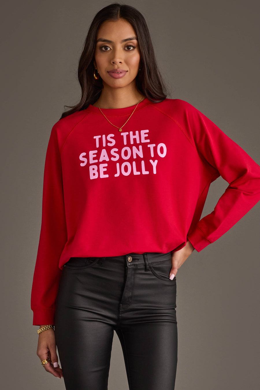 A person wears a red Tis The Season To Be Jolly Sweatshirt with pink lettering, paired with black pants.