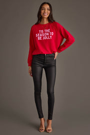 A woman wearing a red Panache Apparel Collection Tis The Season To Be Jolly Sweatshirt with white letters, black skinny jeans, and heeled sandals.