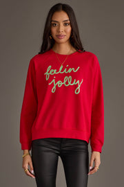 A person wearing an oversized red Feelin Jolly Sweatshirt by Panache Apparel Collection with the text embroidered in green script across the chest.