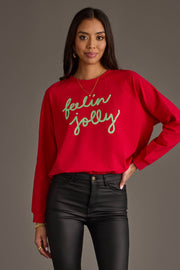 A person wears an oversized red Feelin Jolly Sweatshirt by Panache Apparel Collection with Feelin Jolly written in green cursive text across the chest.