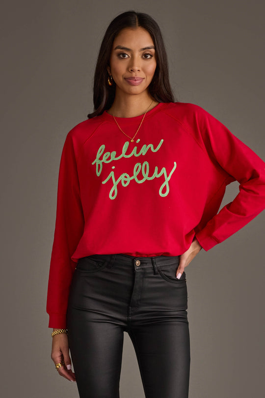 A person wears an oversized red Feelin Jolly Sweatshirt by Panache Apparel Collection with Feelin Jolly written in green cursive text across the chest.