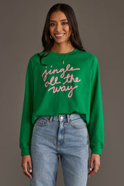 A woman wears a green Jingle All The Way Sweatshirt with the text embroidered in pink script across the front.