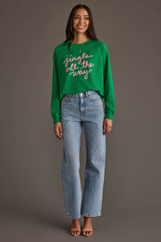 A woman wearing a Green Jingle All The Way Sweatshirt with pink script, paired with light-wash straight-leg jeans.