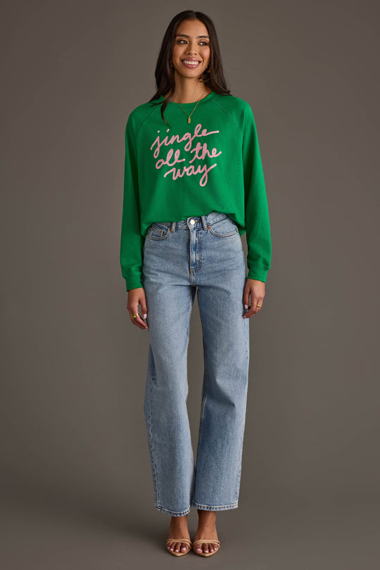 A woman wearing a Green Jingle All The Way Sweatshirt with pink script, paired with light-wash straight-leg jeans.