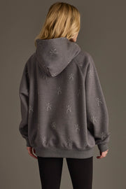 A person with long blonde hair, seen from behind, is wearing the Panache Apparel Collection’s Dancing Skeletons Hooded Sweatshirt, a loose gray hoodie featuring small white stick figure designs, paired with black leggings, standing against a plain, neutral background.