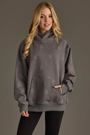 A woman with long blonde hair wears the Panache Apparel Collection’s Dancing Skeletons Hooded Sweatshirt, a loose gray hoodie featuring small stick figure skeleton prints, paired with black leggings, as she stands against a plain brown background.