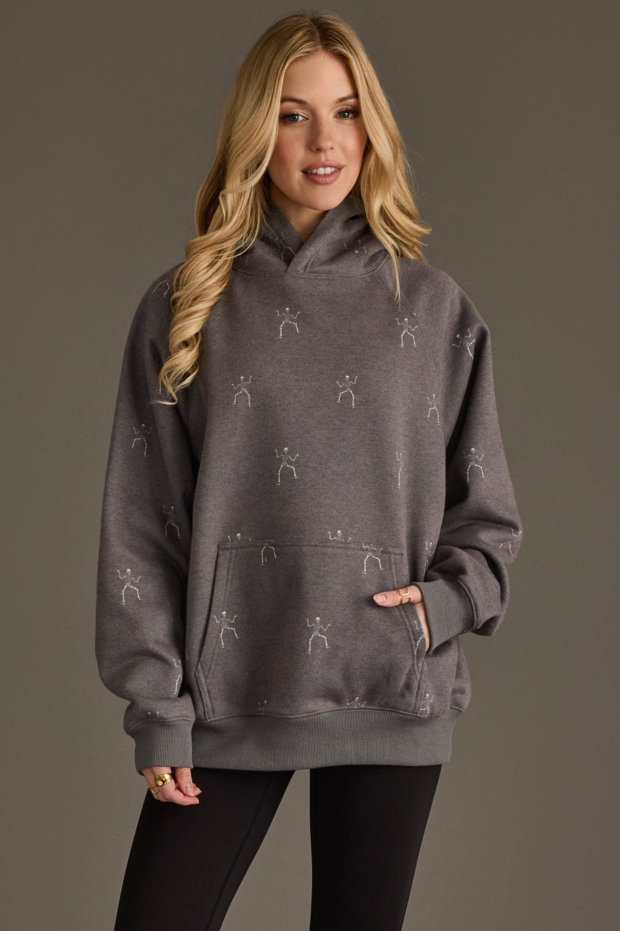 A woman with long blonde hair wears the Panache Apparel Collection’s Dancing Skeletons Hooded Sweatshirt, a loose gray hoodie featuring small stick figure skeleton prints, paired with black leggings, as she stands against a plain brown background.